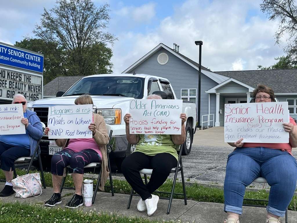 four older persons hold hand written political action informational signs outside Greenville senior center facility.
