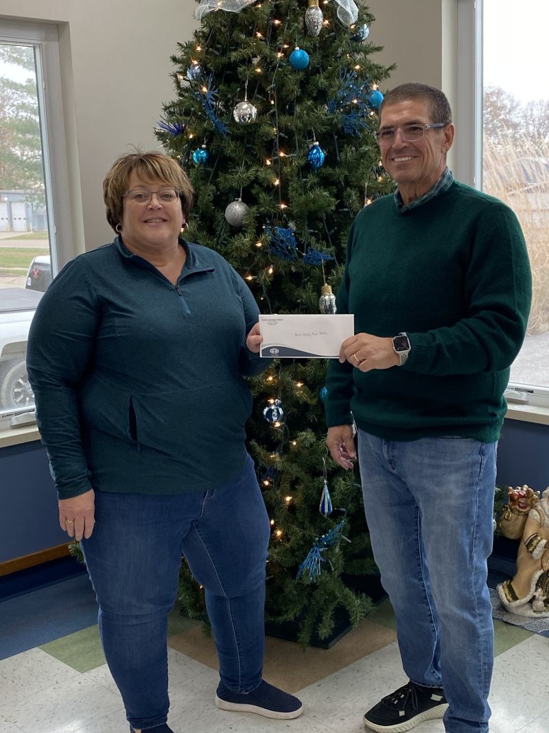 woman and man in green shirts and blue denim jeans hold a check envelope between them, standing in front of evergreen tree decorated with silver ornaments.