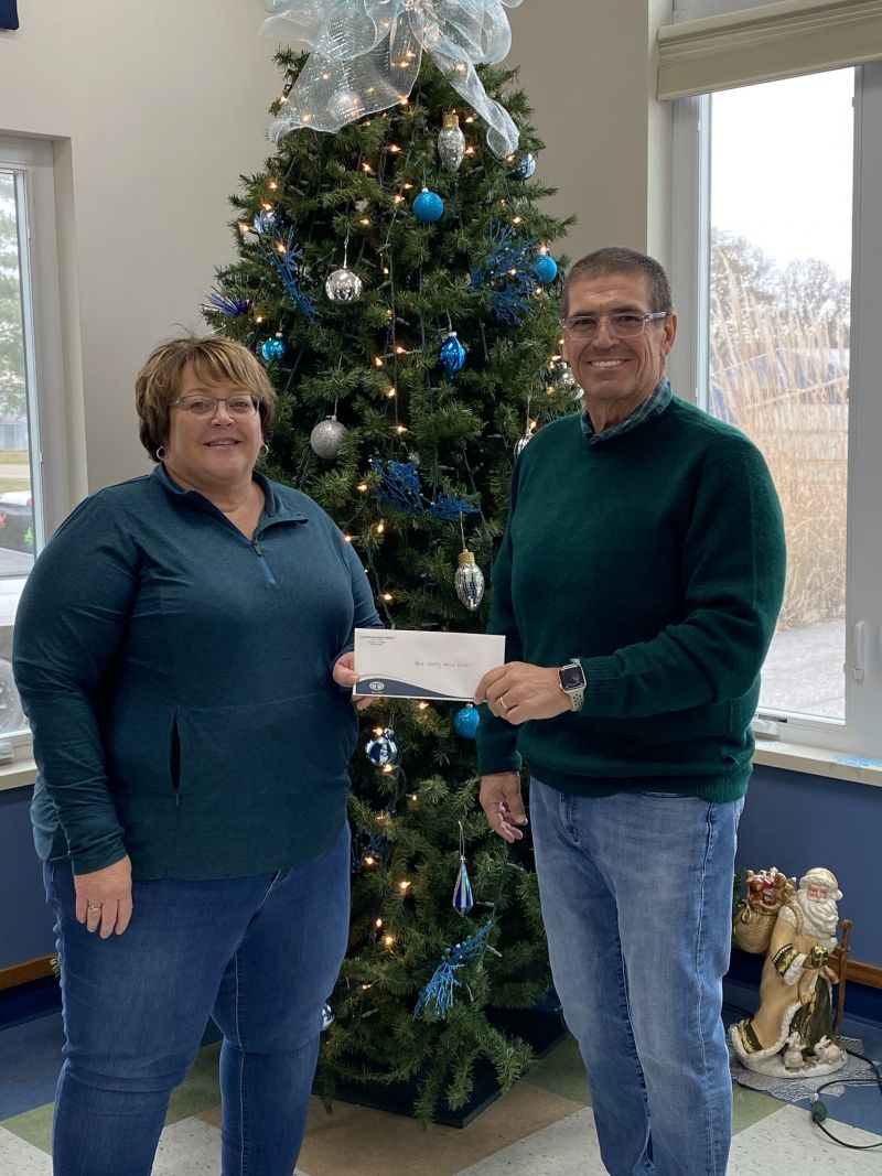 indoors by a window, woman and man in green shirts and blue denim jeans hold a check envelope between them.